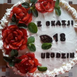 Tort urodzinowy z białym kremem, dekorowany truskawkowymi różami, listkami mięty i czekoladowym napisem 'Z okazji 18 Urodzin', leżący na drewnianej desce.