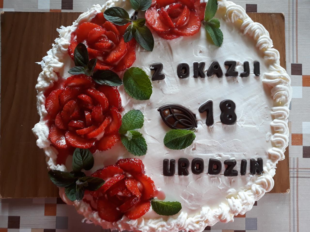 Tort urodzinowy z białym kremem, dekorowany truskawkowymi różami, listkami mięty i czekoladowym napisem 'Z okazji 18 Urodzin', leżący na drewnianej desce.