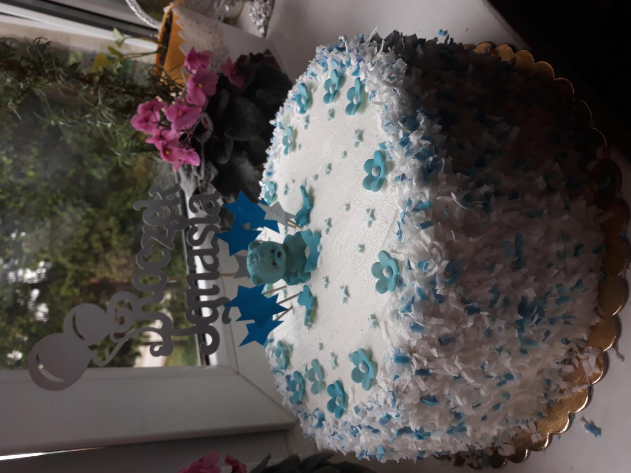 Okrągły tort z białym kremem, posypką kokosową i błękitnymi kwiatkami, ozdobiony niebieskim misiem i gwiazdkami oraz napisem 'Roczek Donatasa'.