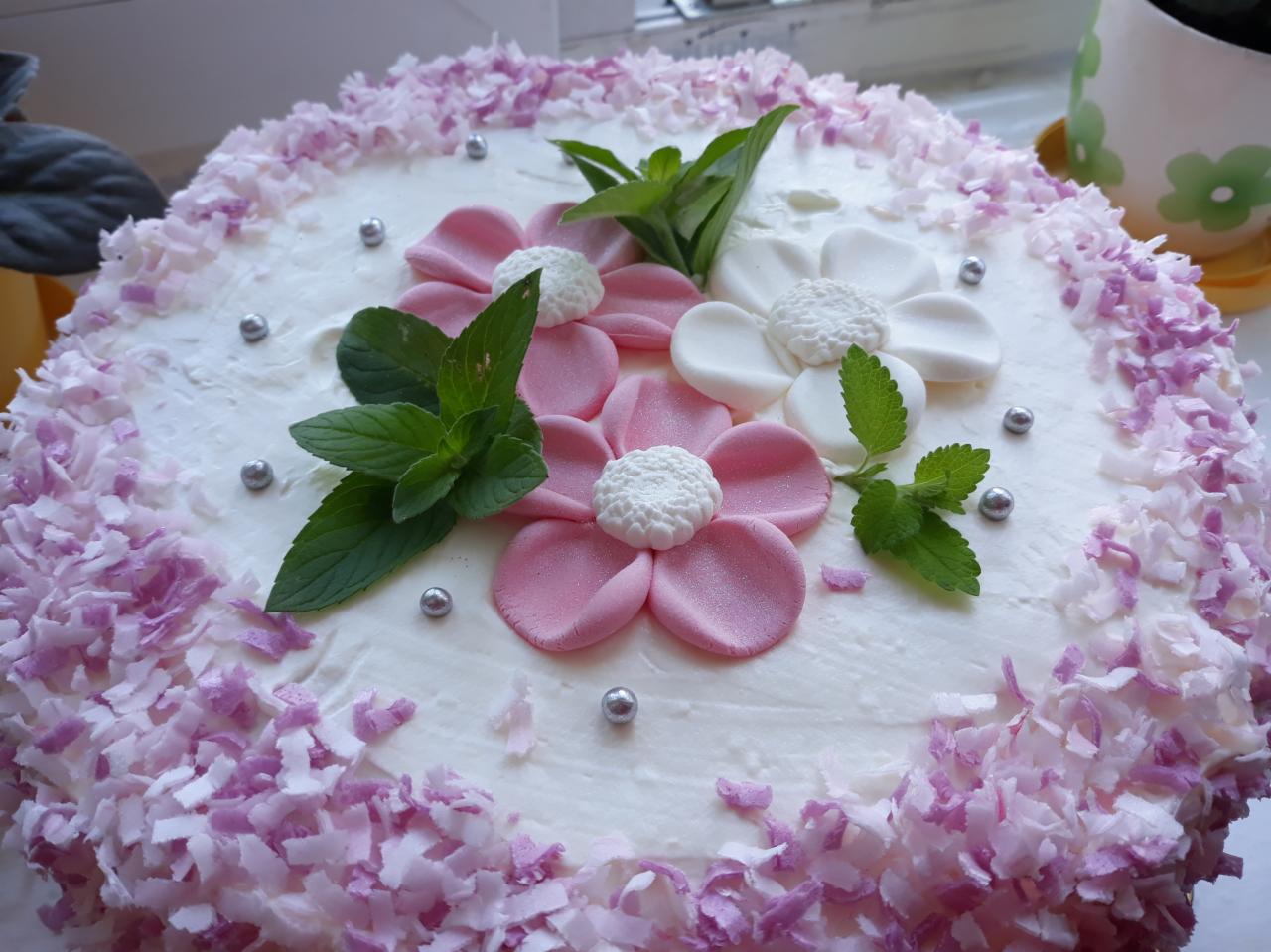 Tort udekorowany białym kremem, różowo-białymi kwiatami z lukru, świeżymi listkami mięty i posypką z wiórków kokosowych barwionych na fioletowo, ozdobiony srebrnymi kuleczkami.
