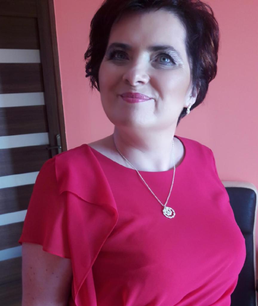 Portret kobiety w czerwonej sukience, z krótkimi ciemnymi włosami i srebrnym naszyjnikiem, uśmiechającej się i patrzącej w górę, na tle różowej ściany.