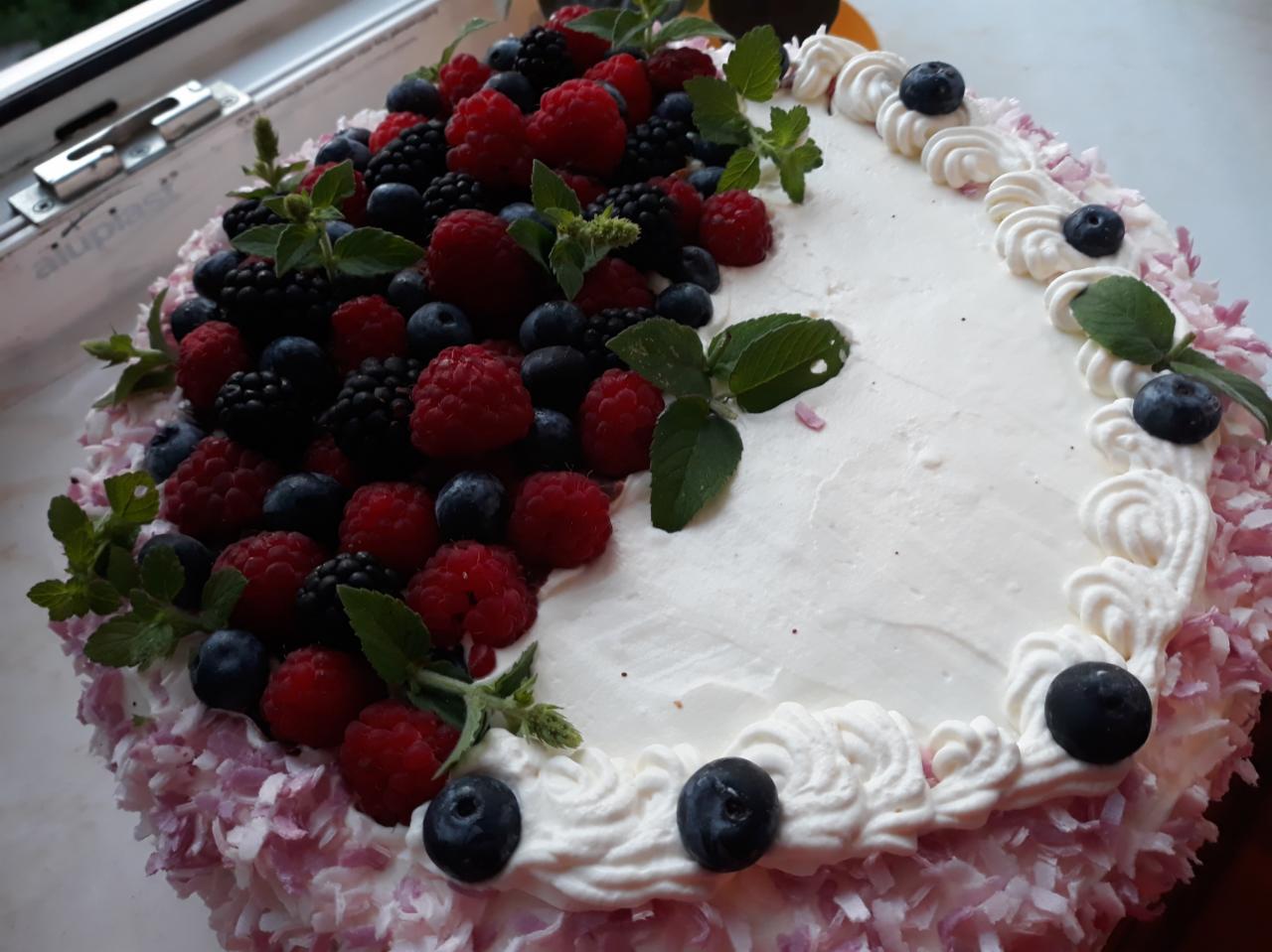 Okrągły tort z białym kremem, udekorowany świeżymi malinami, jeżynami, borówkami i listkami mięty, z obrzeżem z białych rozetek kremu i płatków kokosowych barwionych na delikatny róż.