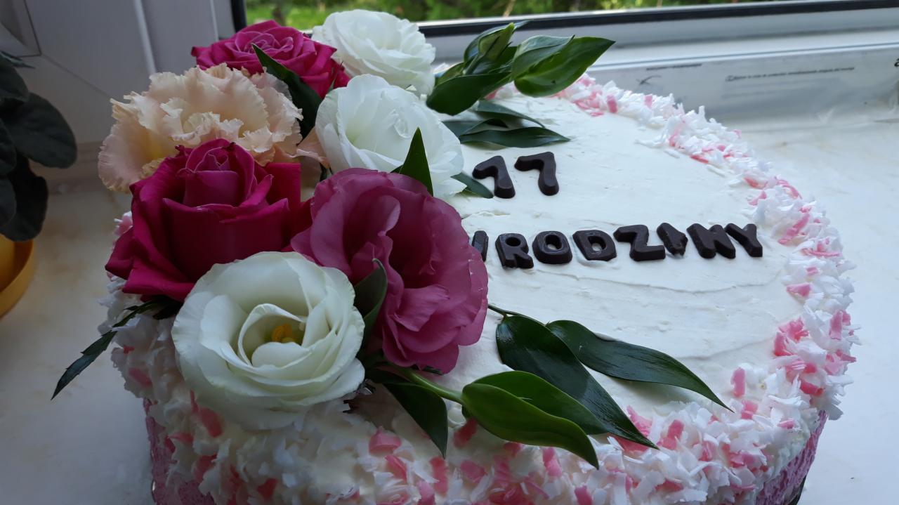Tort urodzinowy z białym kremem, dekorowany świeżymi różami w odcieniach różu, bieli i kremu, z napisem '17 Urodziny' wykonanym z ciemnej czekolady, otoczony wiórkami kokosowymi z różowymi akcentami.