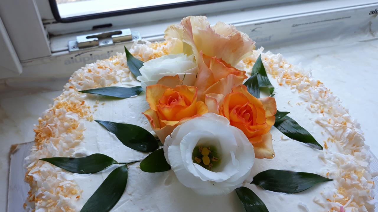 Tort z białym kremem, posypany wiórkami kokosowymi z pomarańczowymi akcentami i udekorowany kompozycją z białych i pomarańczowych róż oraz zielonych liści, sfotografowany w pobliżu okna.