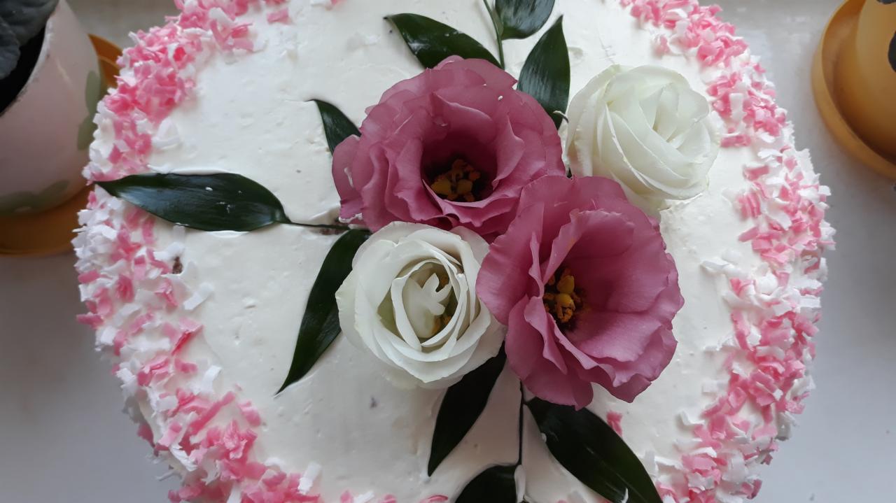 Tort z białym kremem, obsypany różowo-białymi wiórkami kokosowymi, udekorowany trzema kwiatami (dwoma różowymi i jednym białym) oraz zielonymi liśćmi, widok z góry.