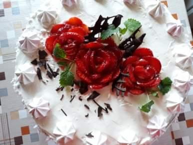 Tort z białym kremem, dekorowany rozetami z truskawek, bezikami i czekoladowymi wiórkami, ułożony na stole z wzorzystym obrusem.