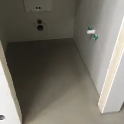 Świeżo wylana, gładka, szara posadzka w pomieszczeniu z widocznymi instalacjami hydraulicznymi i elektrycznymi w ścianach, przygotowana do dalszych prac wykończeniowych.