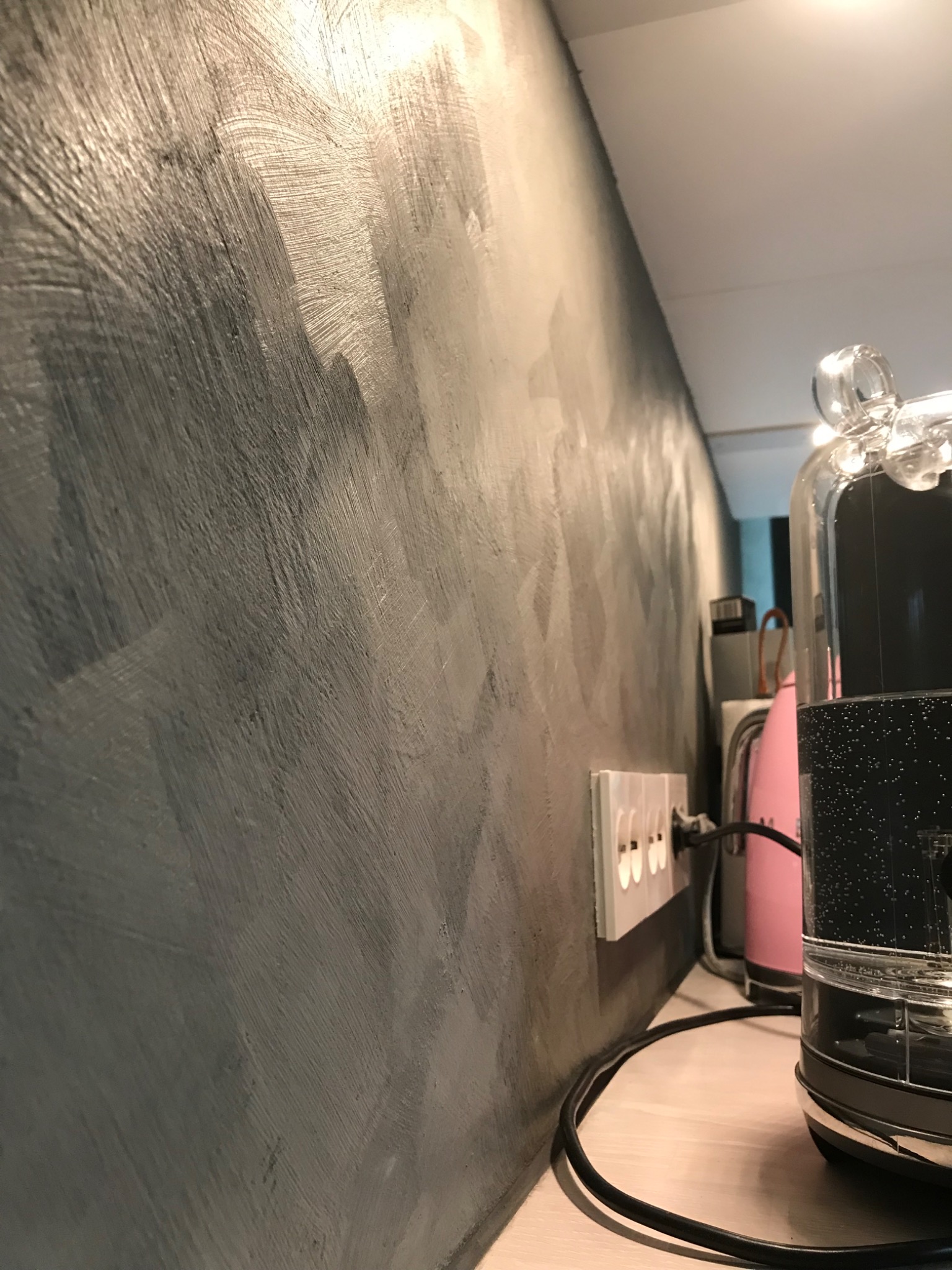 Szara ściana z efektem dekoracyjnym przypominającym przecierkę, widoczne gniazdko elektryczne i fragmenty urządzeń AGD na blacie.