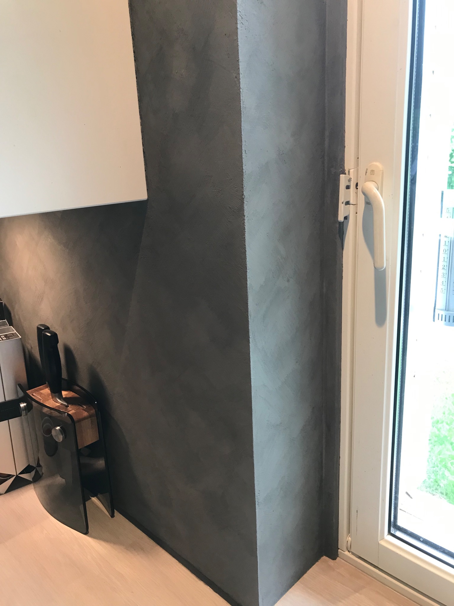 Szara ściana wykończona w stylu betonu architektonicznego, sąsiadująca z białym oknem i fragmentem białej szafki, na jasnej drewnianej podłodze.