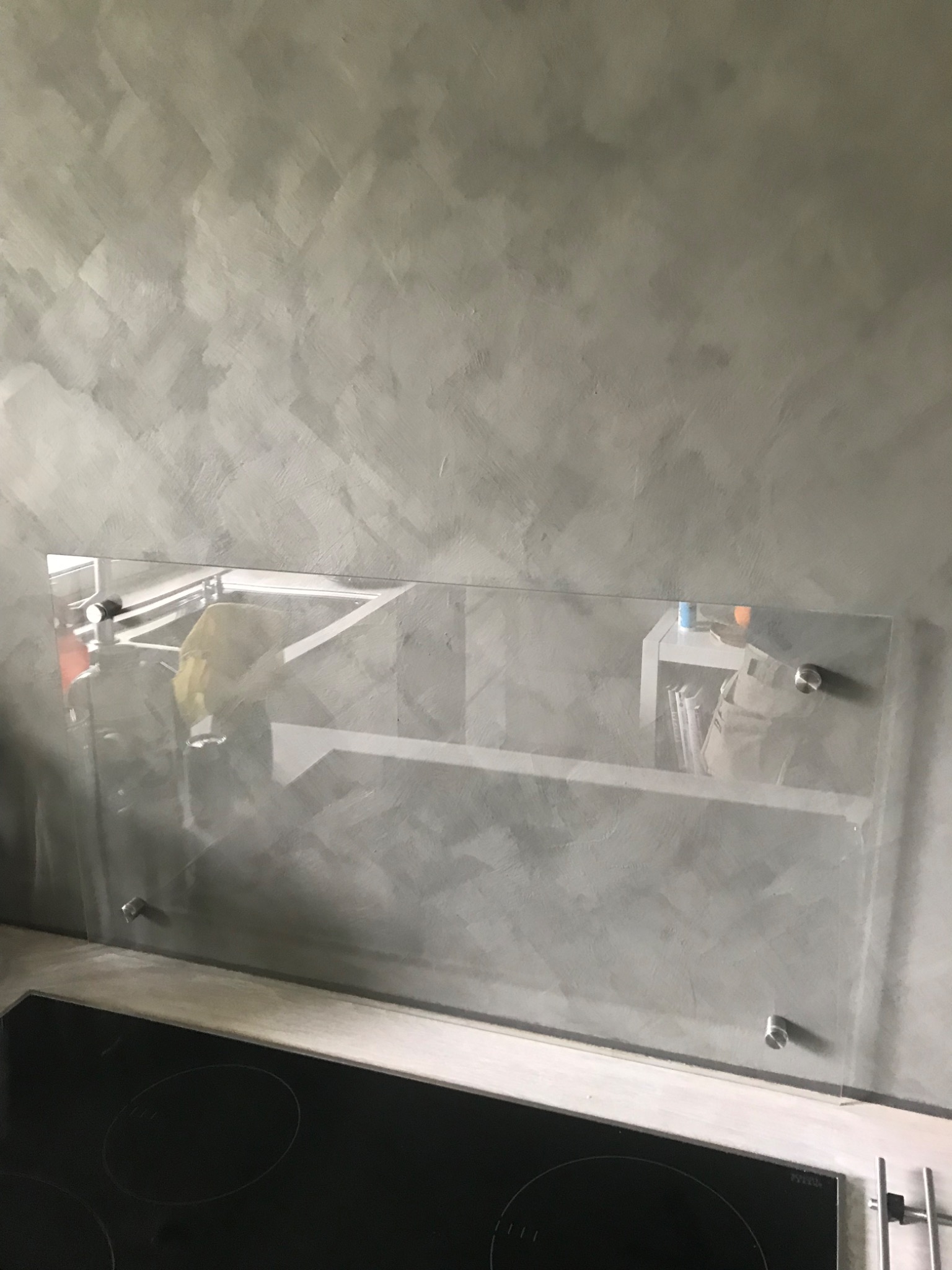 Ściana w kuchni pokryta szarym mikrocementem z zamontowanym szklanym panelem nad płytą indukcyjną.