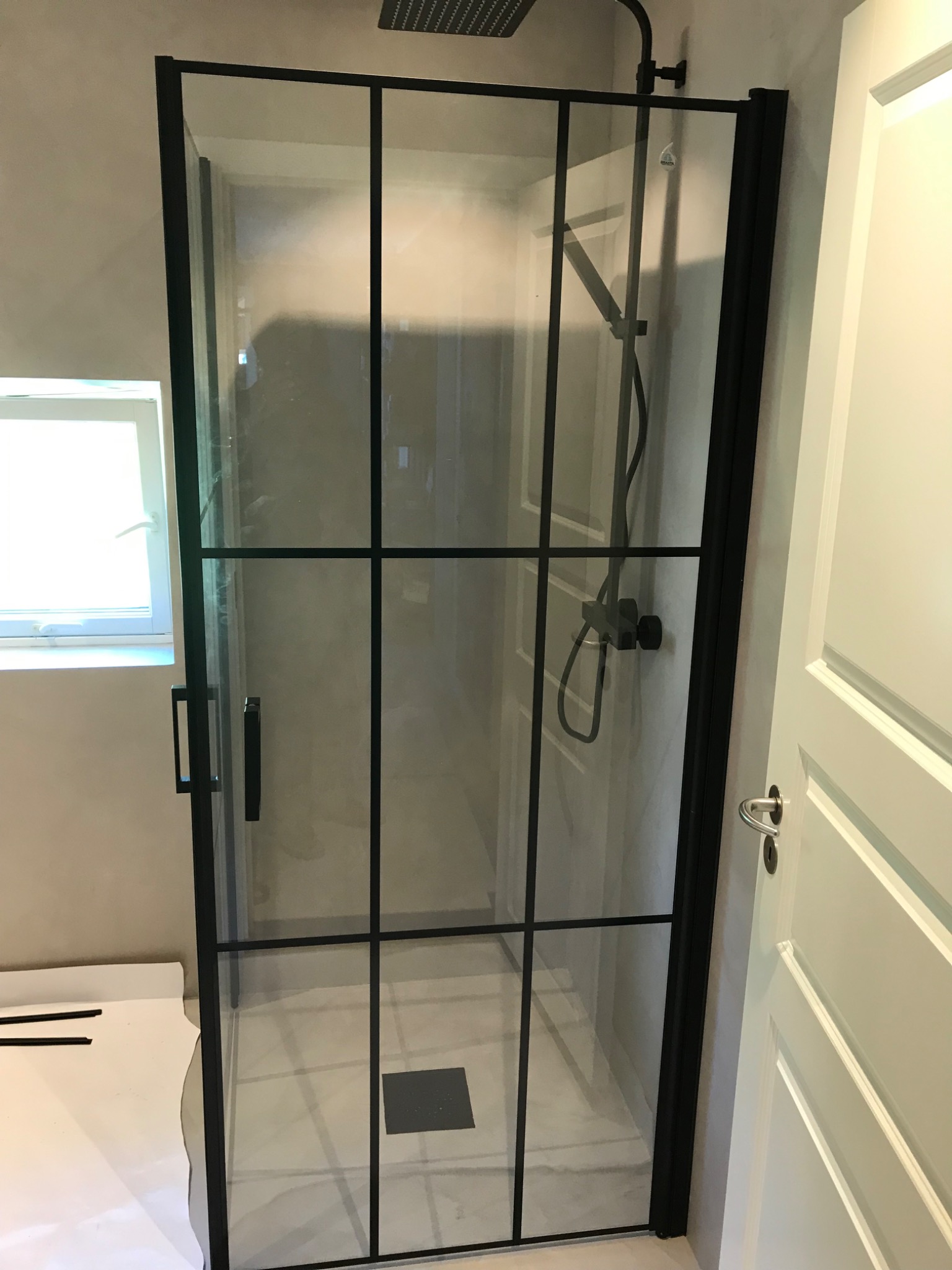 Nowoczesna kabina prysznicowa z czarnymi profilami i szklanymi ściankami, widoczny deszczownica i bateria, jasne płytki podłogowe, obok białe drzwi i okno.