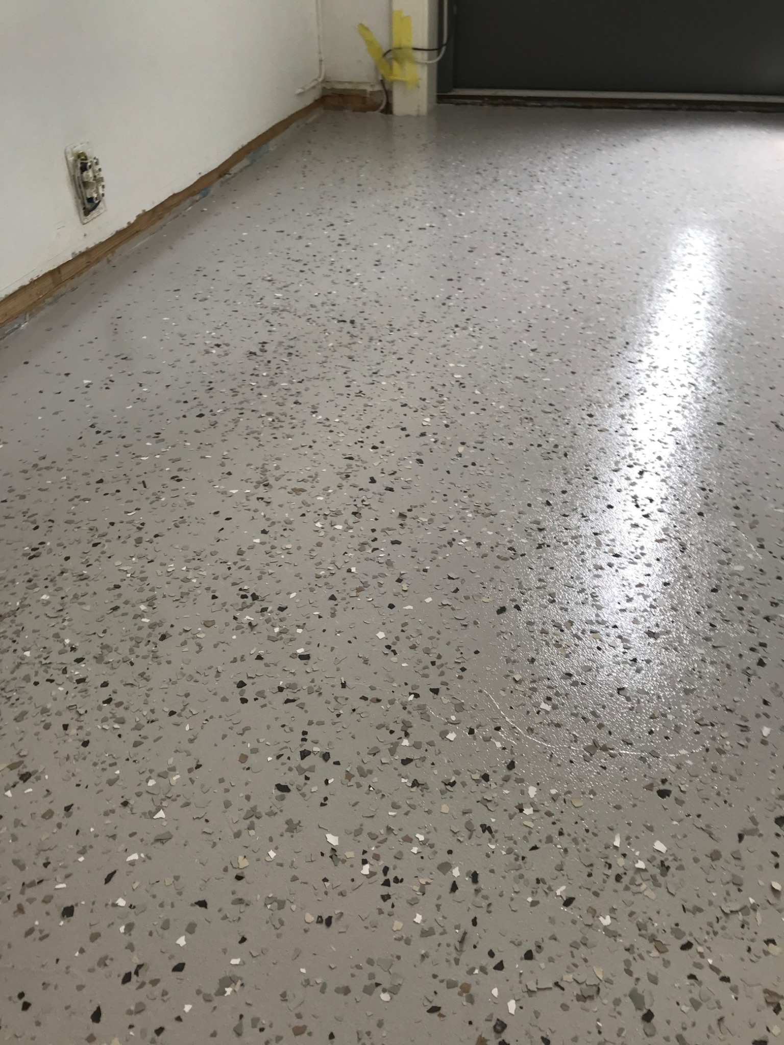 Jasnoszara, błyszcząca podłoga z drobnymi, wielobarwnymi płatkami, widoczna przy ścianie z niedokończonym wykończeniem i gniazdkiem elektrycznym.