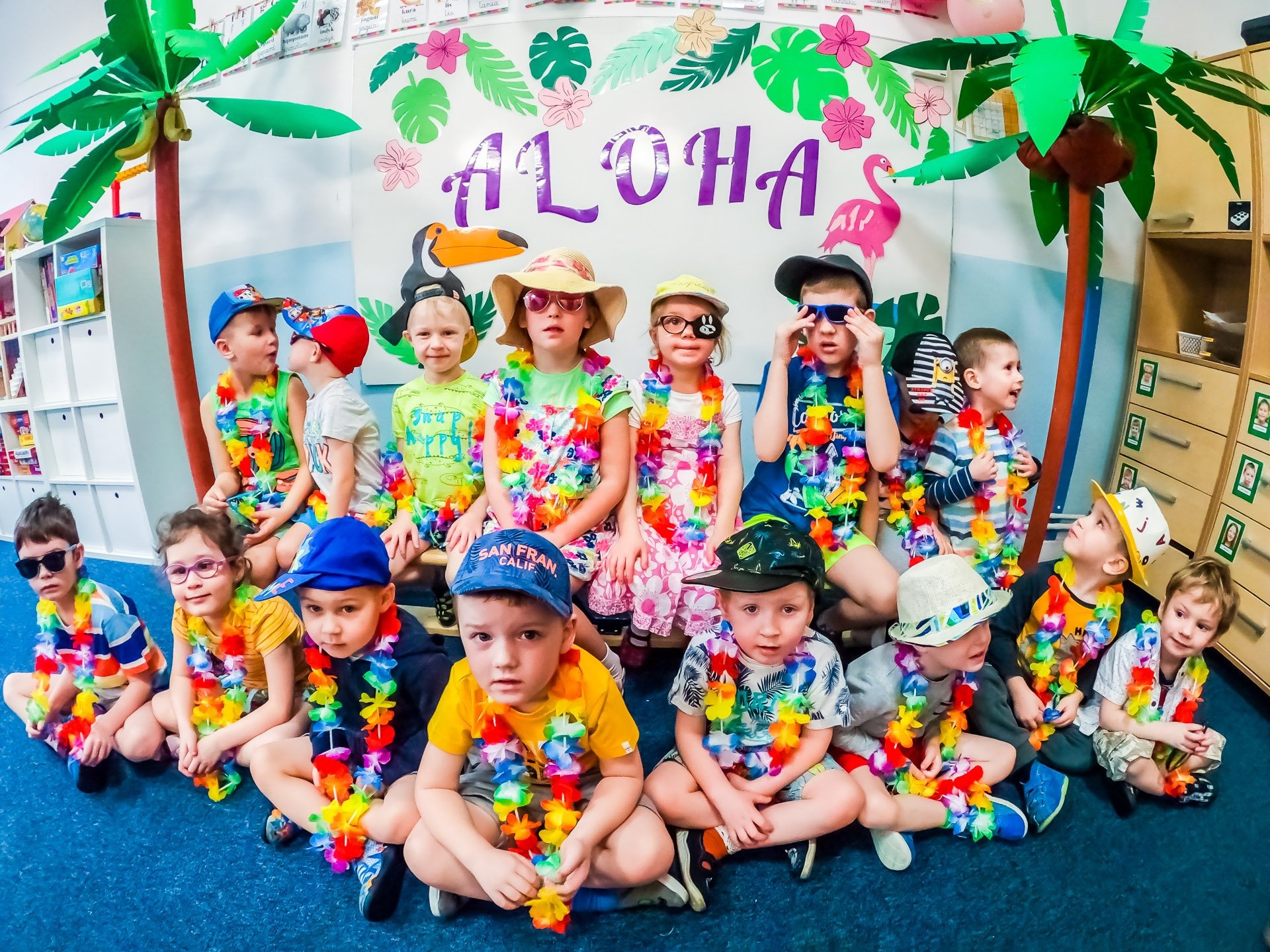 Grupa dzieci w hawajskich wieńcach siedzi na tle dekoracji z palmami i napisem Aloha, niektóre dzieci mają okulary przeciwsłoneczne i kapelusze.