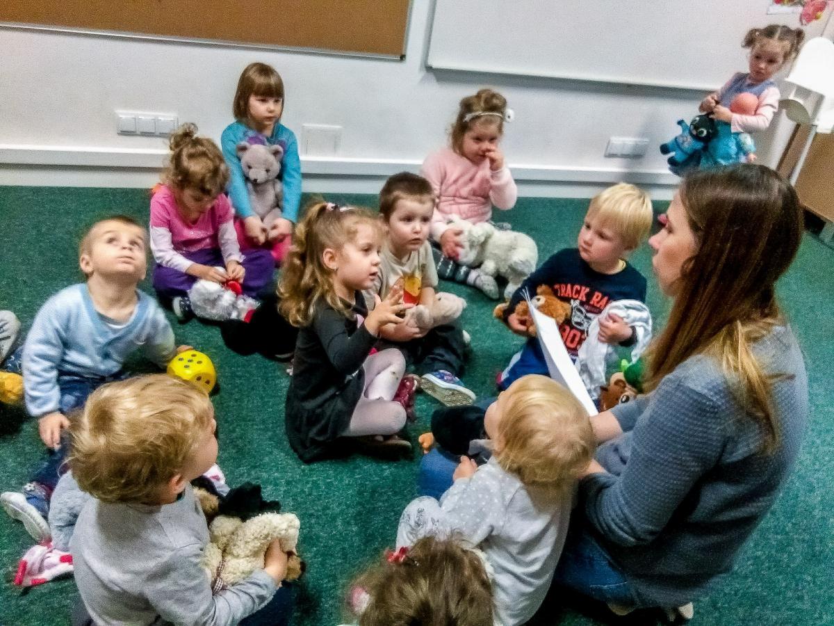 Grupa dzieci siedzi na zielonym dywanie w kręgu, słuchając kobiety czytającej z kartki, każde dziecko trzyma pluszową zabawkę, jedno z dzieci patrzy w górę.