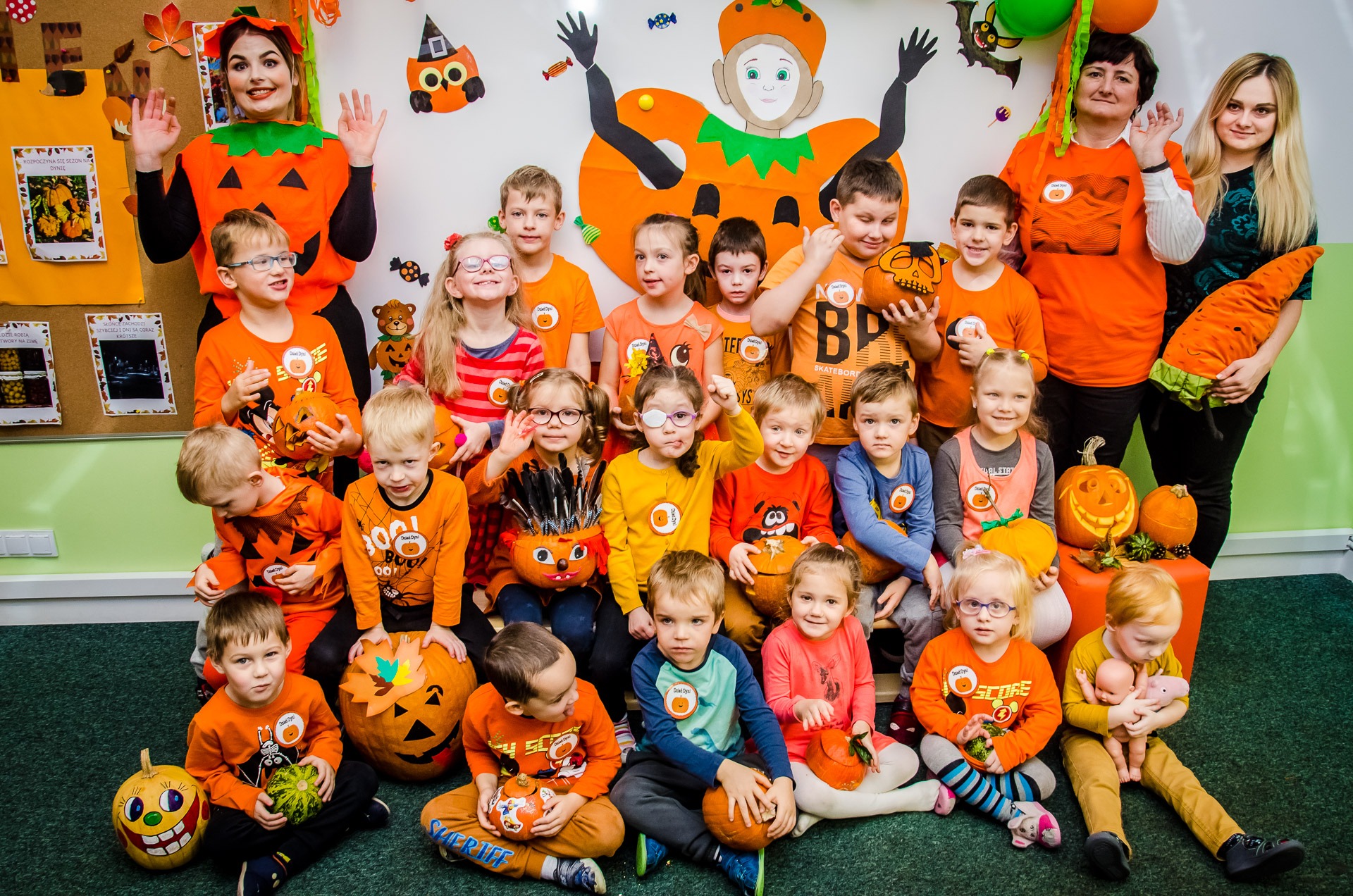 Grupa dzieci w pomarańczowych strojach z motywami Halloween, trzymających dynie, pozuje do zdjęcia z dwiema opiekunkami w przebraniach dyni, w tle dekoracje halloweenowe na ścianie i tablicy.