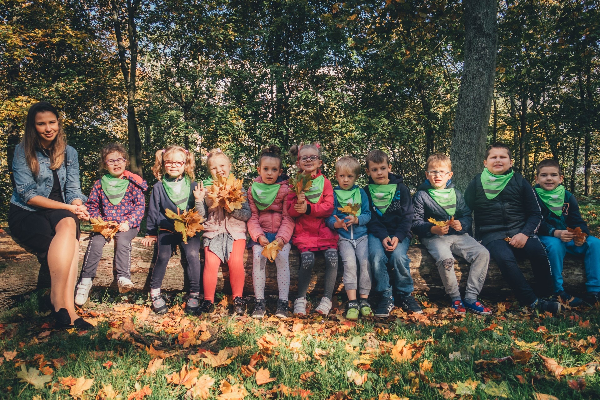 Grupa dzieci w zielonych chustach i wychowawczyni siedzą na powalonym pniu drzewa w jesiennym lesie, trzymając w dłoniach liście.