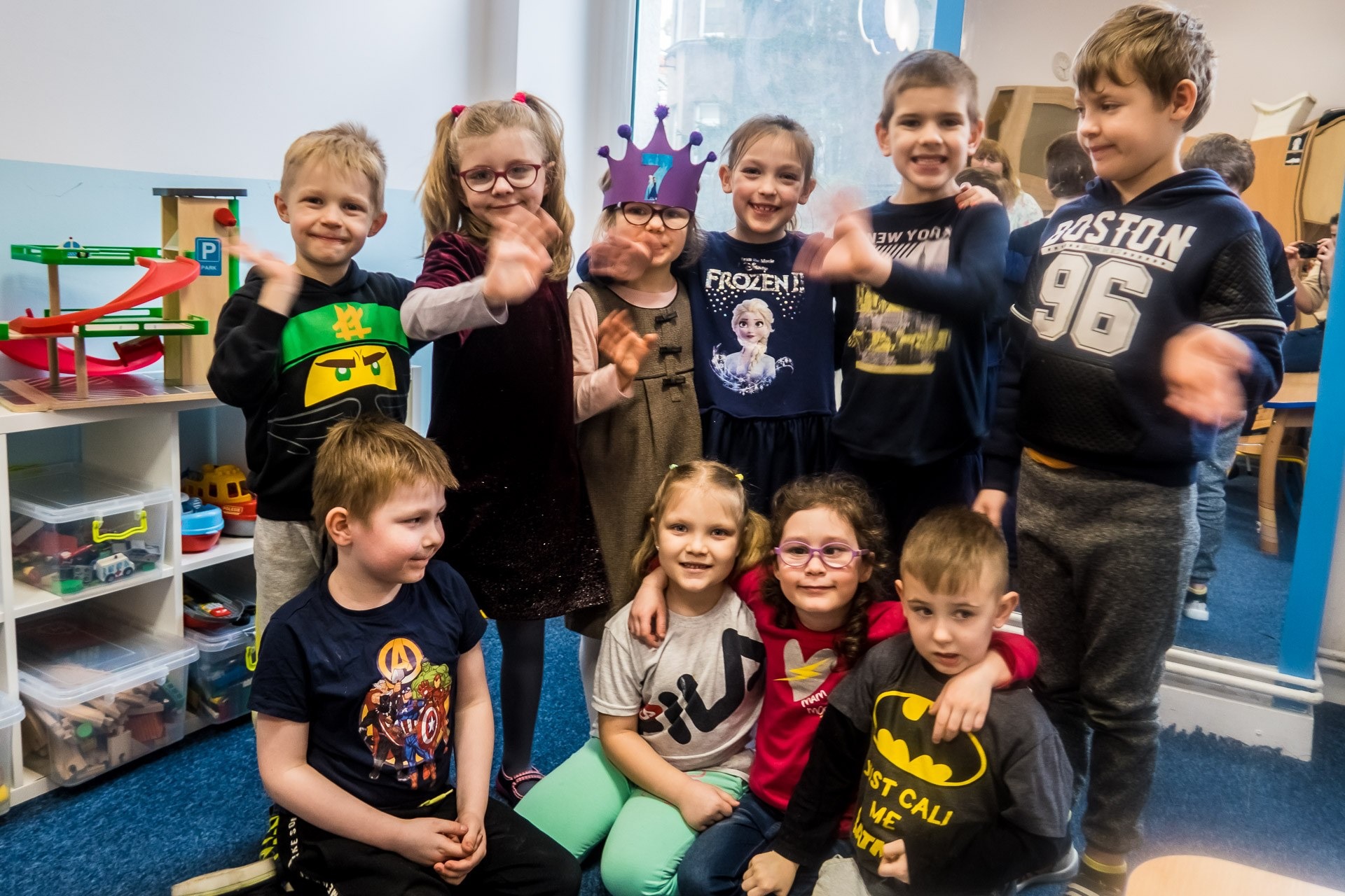 Grupa uśmiechniętych dzieci w różnym wieku, niektóre w okularach i kostiumach, pozują w sali zabaw z półkami pełnymi zabawek w tle, wesoła atmosfera.