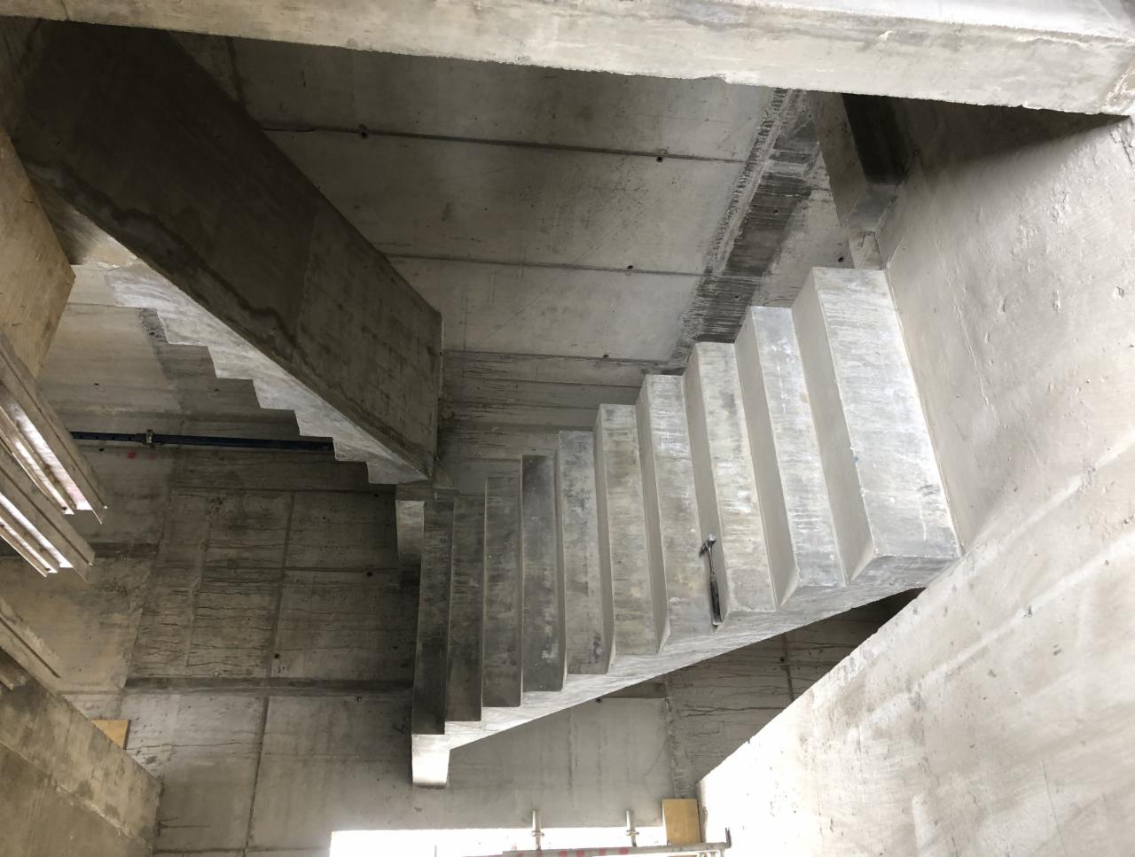 Surowe betonowe schody w trakcie budowy, widok z dołu, szalunki częściowo zdemontowane, widoczne ślady po deskach na ścianach.