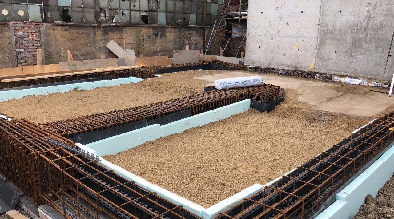 Przygotowanie terenu pod wylewkę fundamentów: szalunek zbrojeniowy, termoizolacja ze styropianu i podsypka piaskowa, na tle starego budynku przemysłowego z powybijanymi oknami.