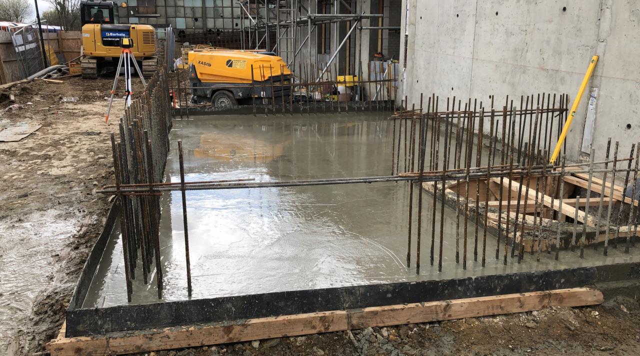 Zalany betonem szalunek z wystającymi prętami zbrojeniowymi, w tle żółty kompresor i niwelator na placu budowy.