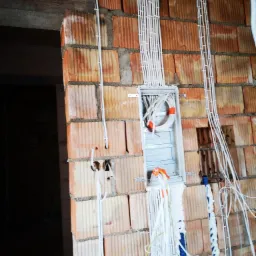 Ściana z cegły dziurawki w trakcie montażu instalacji elektrycznej, widoczne kable prowadzone w peszlach i puszka instalacyjna z zapasem przewodów, pianka montażowa uszczelniająca przejścia.