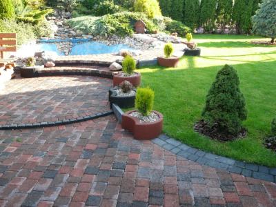 Ułożona kostka brukowa w odcieniach czerwieni i brązu tworząca ścieżkę w ogrodzie z oczkiem wodnym i roślinami w donicach.