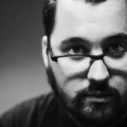 Portret w czerni i bieli: Mężczyzna z brodą i okularami, zamyślone spojrzenie, oświetlenie z lewej strony tworzy wyraźne cienie.