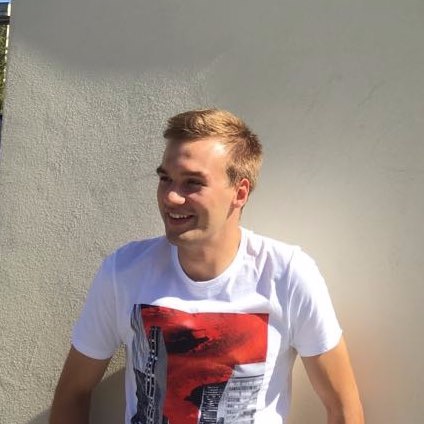 Uśmiechnięty młody mężczyzna w białej koszulce z motywem miejskim na tle jasnej ściany.