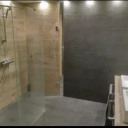 Nowoczesna łazienka z kabiną prysznicową ze szklanymi drzwiami, drewnopodobnymi płytkami ściennymi i ciemnoszarymi płytkami na ścianie obok umywalki.