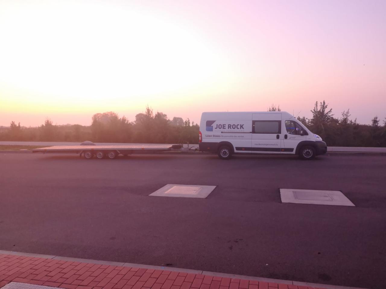 Biały bus marki Joe Rock z przyczepą na tle wschodzącego słońca. Transport pojazdów na lawecie. Asfaltowa droga z elementami infrastruktury.