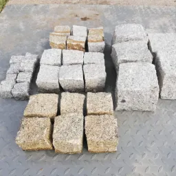 Różne rozmiary kostki granitowej w odcieniach szarości i beżu ułożone na metalowej, ryflowanej powierzchni.