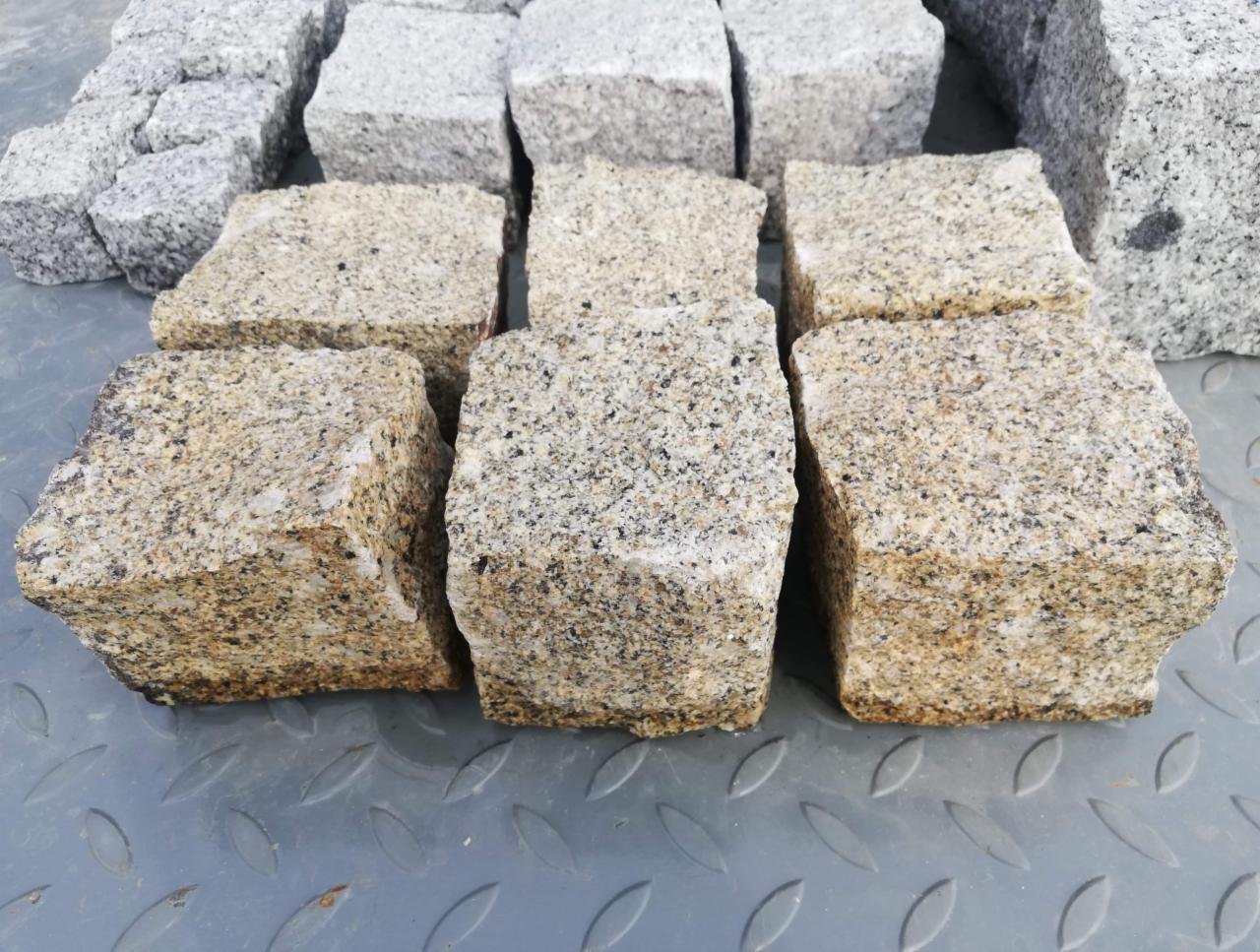 Rząd ułożonych kostek z jasnego, ziarnistego granitu na metalowej powierzchni z tłoczeniami, widoczne też szare kostki w tle.