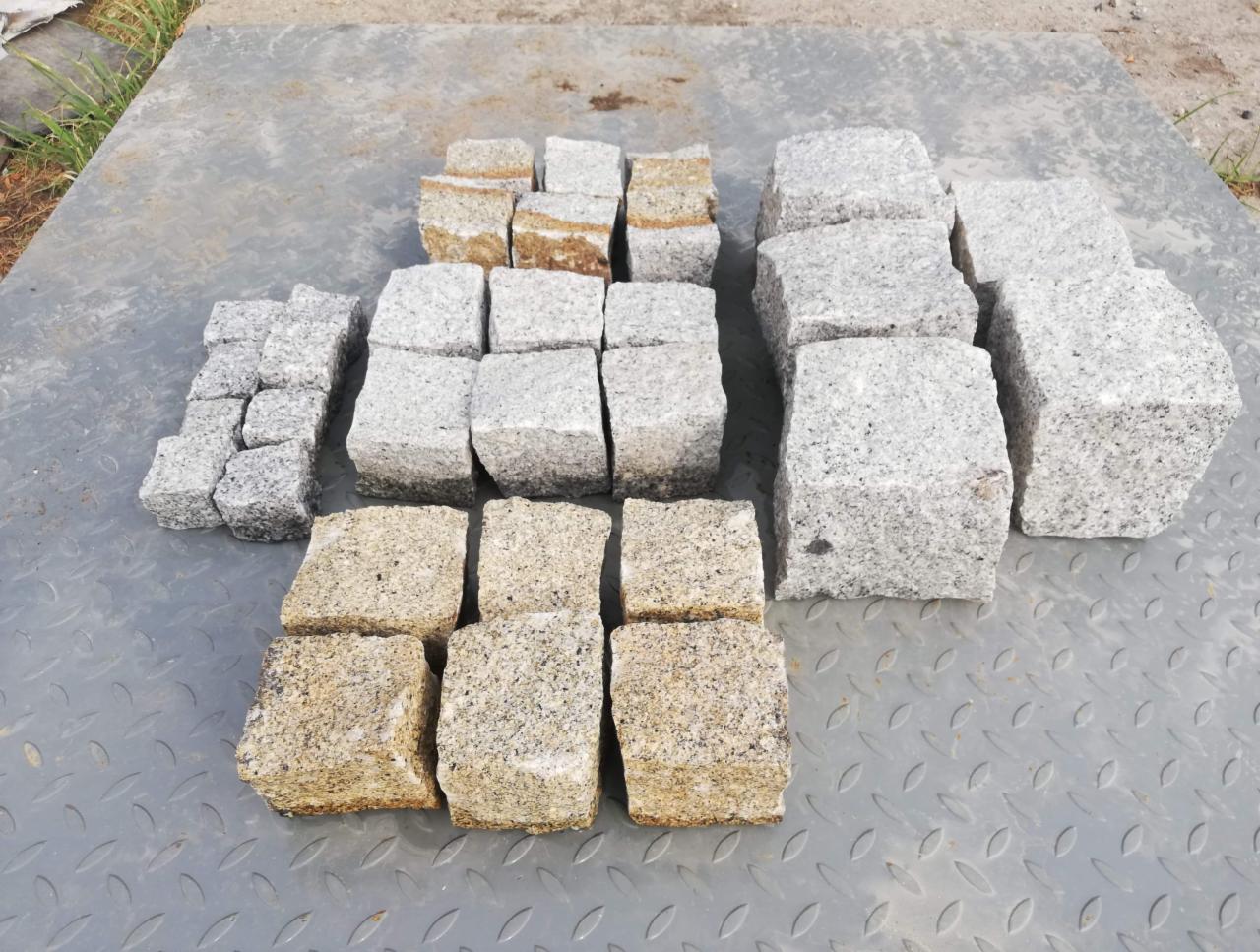 Różne rozmiary kostki granitowej w odcieniach szarości i beżu ułożone na metalowej, ryflowanej powierzchni.