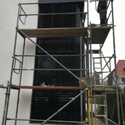 Pracownik na rusztowaniu montuje zewnętrzne, poziome żaluzje okienne w nowoczesnym budynku z białą elewacją.