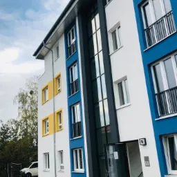 Nowoczesny budynek wielorodzinny z kolorową elewacją (biały, niebieski, żółty), z oknami z czarnymi balustradami i szklanymi drzwiami wejściowymi z numerem 15, widok z zewnątrz.