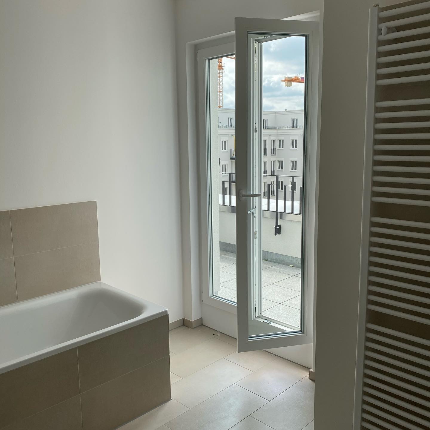 Jasna łazienka z otwartymi białymi drzwiami balkonowymi, widok na balkon i budynek w budowie z żurawiem.