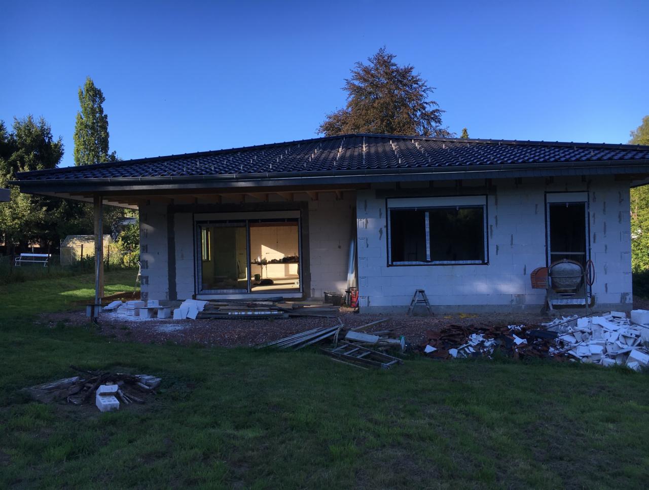 Budowa domu jednorodzinnego w stanie surowym otwartym, widoczne otwory okienne i drzwiowe bez stolarki, na trawniku gruz i materiały budowlane, betoniarka.