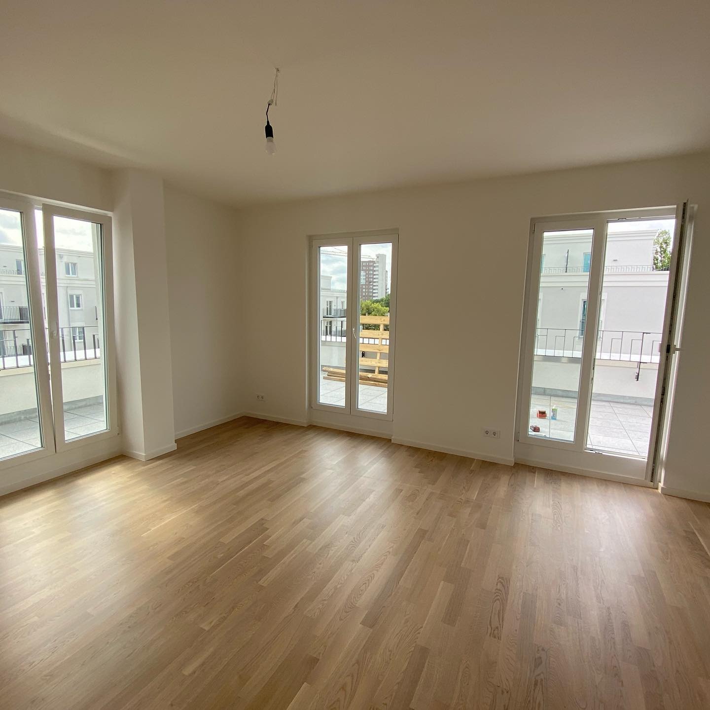 Jasne, puste wnętrze z trzema białymi oknami balkonowymi, parkiet na podłodze, widok na balkon i budynki na zewnątrz, pojedyncza żarówka zwisająca z sufitu.