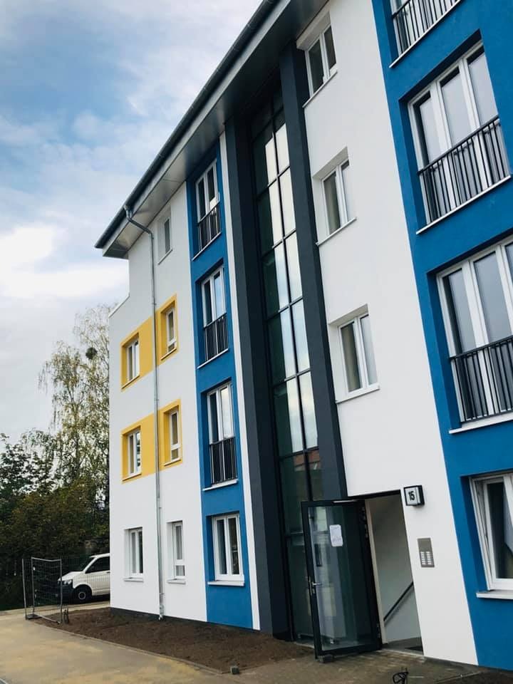 Nowoczesny budynek wielorodzinny z kolorową elewacją (biały, niebieski, żółty), z oknami z czarnymi balustradami i szklanymi drzwiami wejściowymi z numerem 15, widok z zewnątrz.