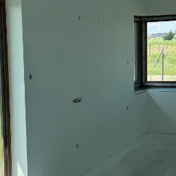 Wnętrze pokoju po malowaniu ścian na jasny odcień szarości, widoczne puszki elektryczne i okna z widokiem na zewnątrz.