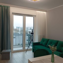 Robert Drwęcki - Jasne wnętrze po wykończeniu: zielona sofa, stół z dekoracją, okno balkonowe z widokiem na miasto. W rogu widoczny statyw fotograficzny.