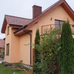 Dom jednorodzinny po malowaniu elewacji na kolor brzoskwiniowy, z widocznym fragmentem dachu z brązowej dach&oacute;wki, balkonem z drewnianą balustradą i ceglanym kominem z daszkiem.