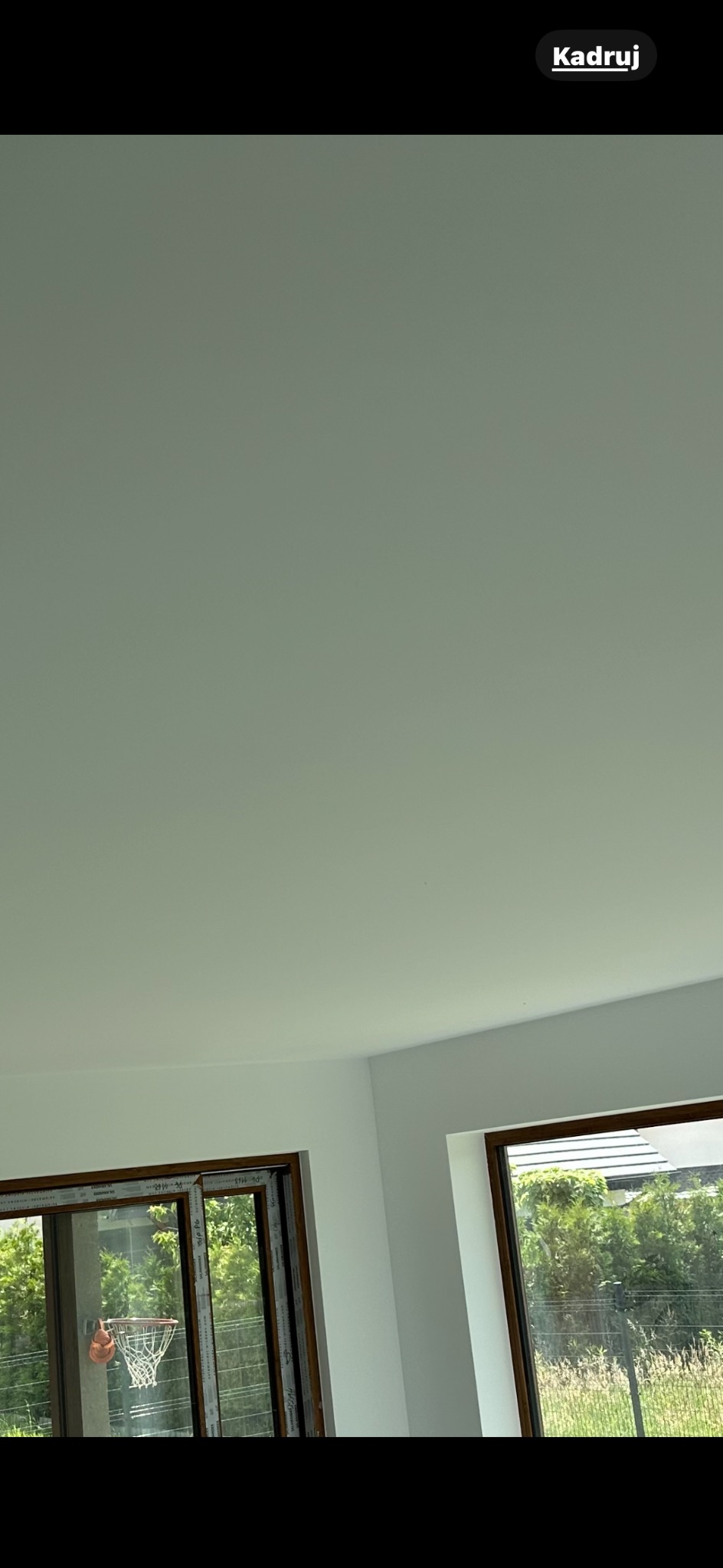 Jasne, świeżo pomalowane wnętrze z dwoma oknami. Przez okna widoczna zieleń i siatka do koszykówki. Minimalistyczny styl wykończenia.