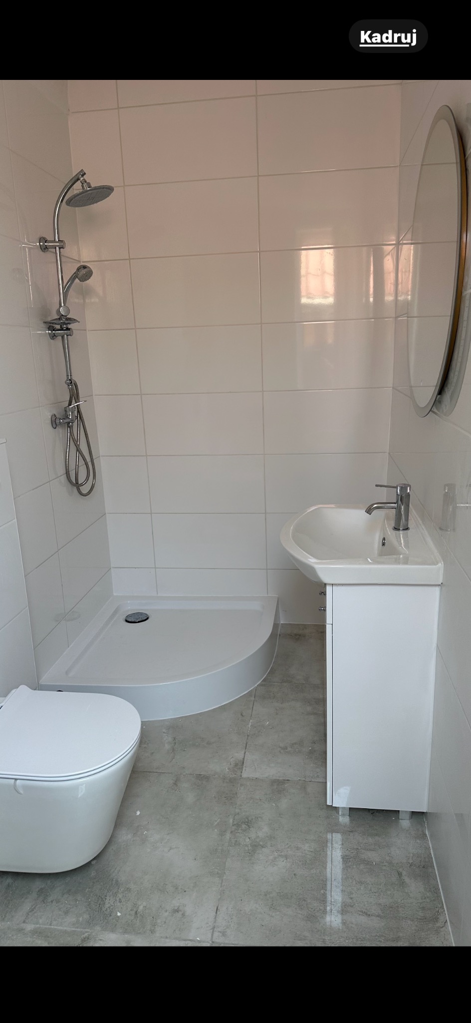 Nowoczesna łazienka po remoncie: białe płytki, kabina prysznicowa z deszczownicą, umywalka z szafką i owalne lustro. Szare płytki podłogowe.