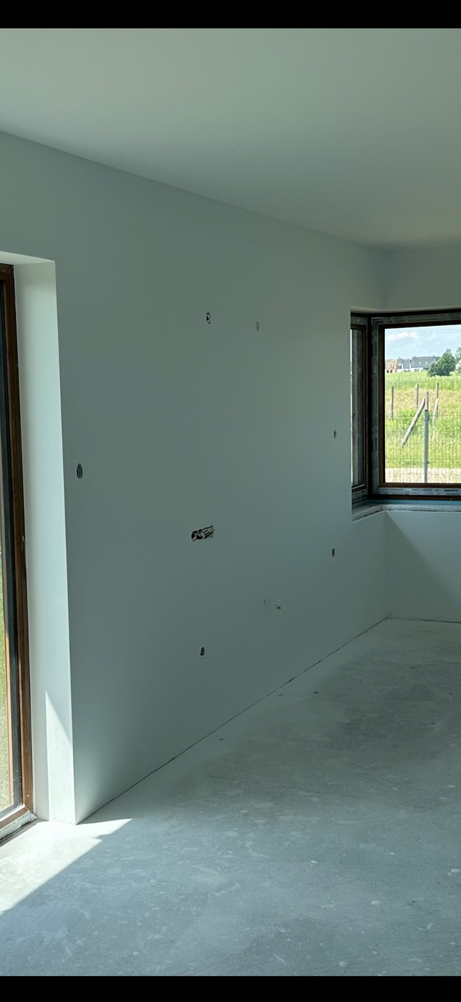 Wnętrze pokoju po malowaniu ścian na jasny odcień szarości, widoczne puszki elektryczne i okna z widokiem na zewnątrz.