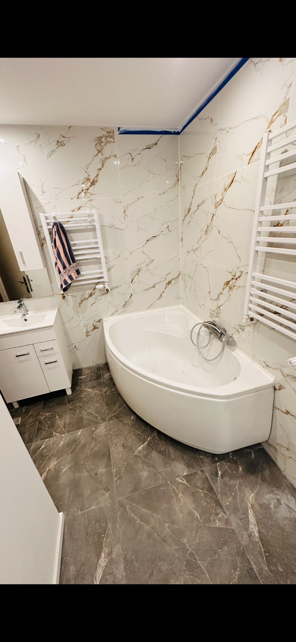 Wykończona łazienka z narożną wanną, marmurowymi płytkami ściennymi i podłogowymi. Widoczna biała szafka z umywalką i grzejnik drabinkowy.