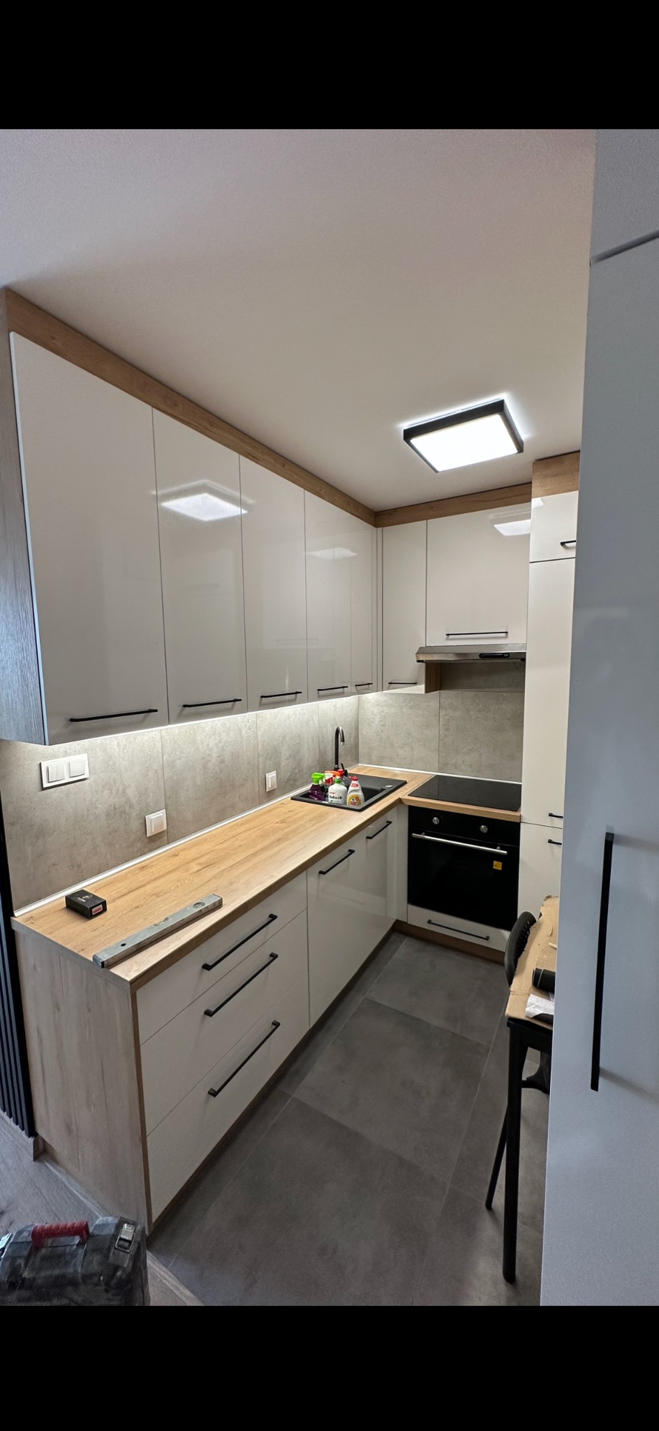Nowoczesna kuchnia z białymi szafkami, drewnianym blatem i szarymi płytkami. Widoczny poziomica na blacie. Ukończone wykończenie wnętrza.