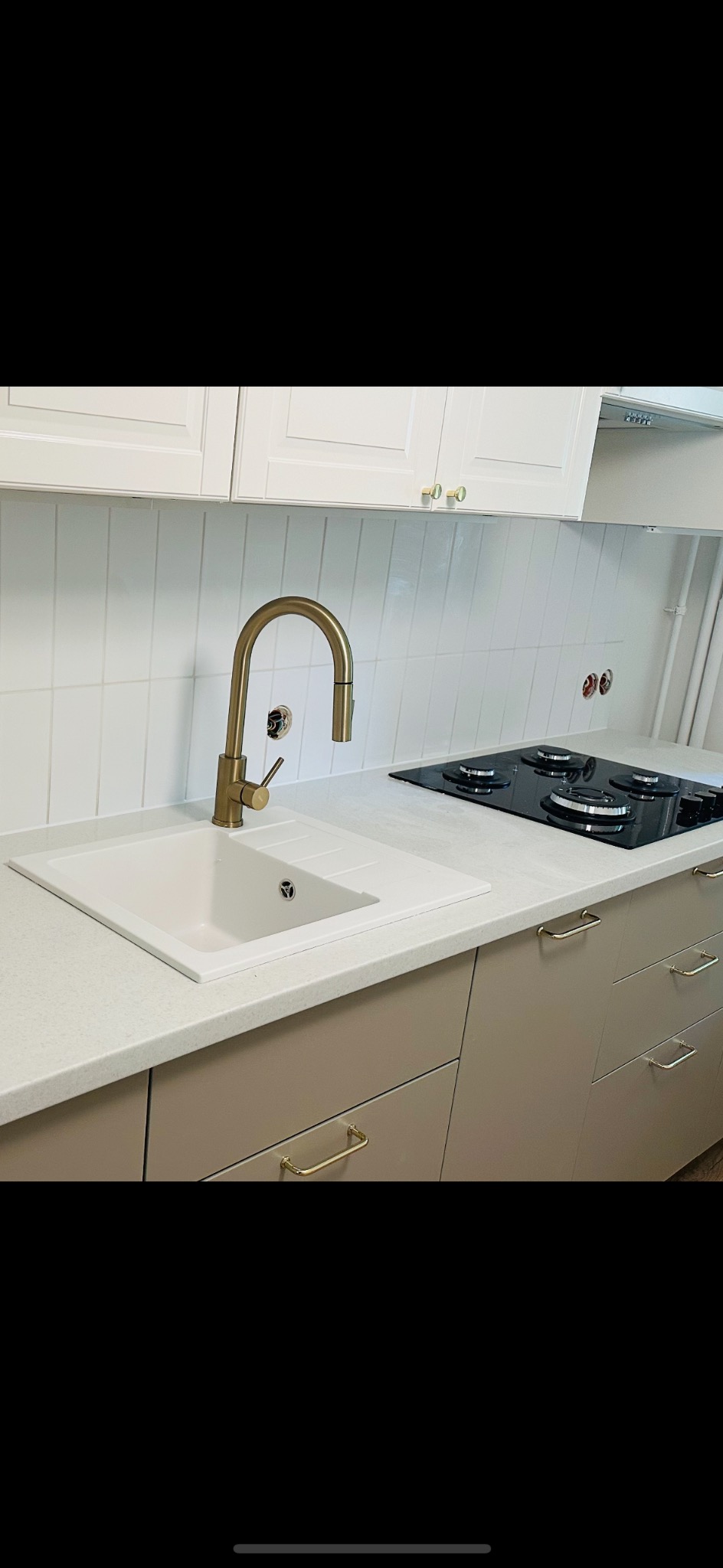 Nowoczesna kuchnia z białymi płytkami, złotą armaturą i beżowymi szafkami. Ujęcie na zlew i płytę grzewczą. Widoczne detale wykończenia.