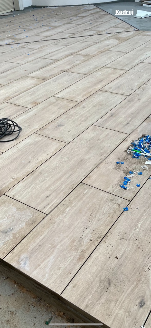 Ułożona podłoga z płytek imitujących drewno, widoczne niebieskie kliny poziomujące, czarny kabel i drobne zabrudzenia. Ujęcie z góry, ukośne.