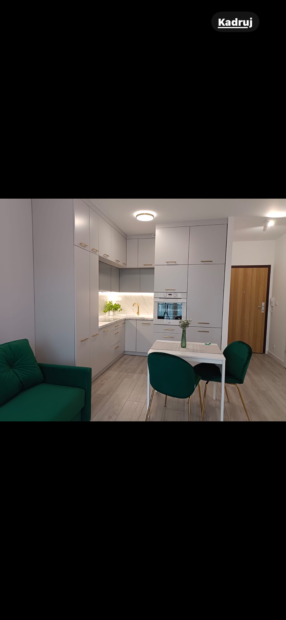 Jasna kuchnia z szarymi szafkami i złotymi uchwytami, uzupełniona zielonymi krzesłami i sofą. Marmurowy blat i złota bateria dodają elegancji.